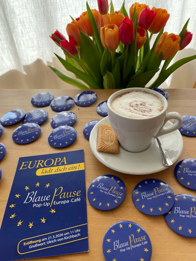 Postkarte und Buttons der Blauen Pause liegen auf einem Tisch, dazu Kaffee und Blumen
