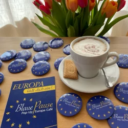 Postkarte und Buttons der Blauen Pause liegen auf einem Tisch, dazu Kaffee und Blumen